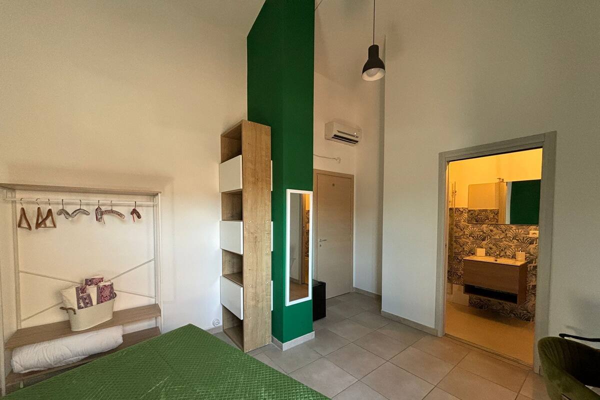 Une chambre avec un mur d’accent vert, un lit recouvert d’une couverture verte, une armoire, un miroir et une salle de bain visible à travers une porte ouverte.