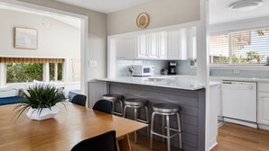 Cozinha moderna com armários brancos, uma ilha central com banquetas e uma área de jantar com mesa e cadeiras de madeira.