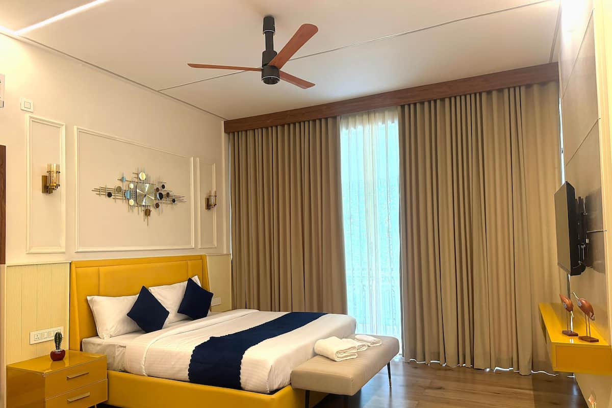 Ein modernes Schlafzimmer mit einem gelben Bett, einem Deckenventilator, einem großen Fenster mit Vorhängen, einer Wanduhr und einem kleinen Nachttisch mit einer Lampe.