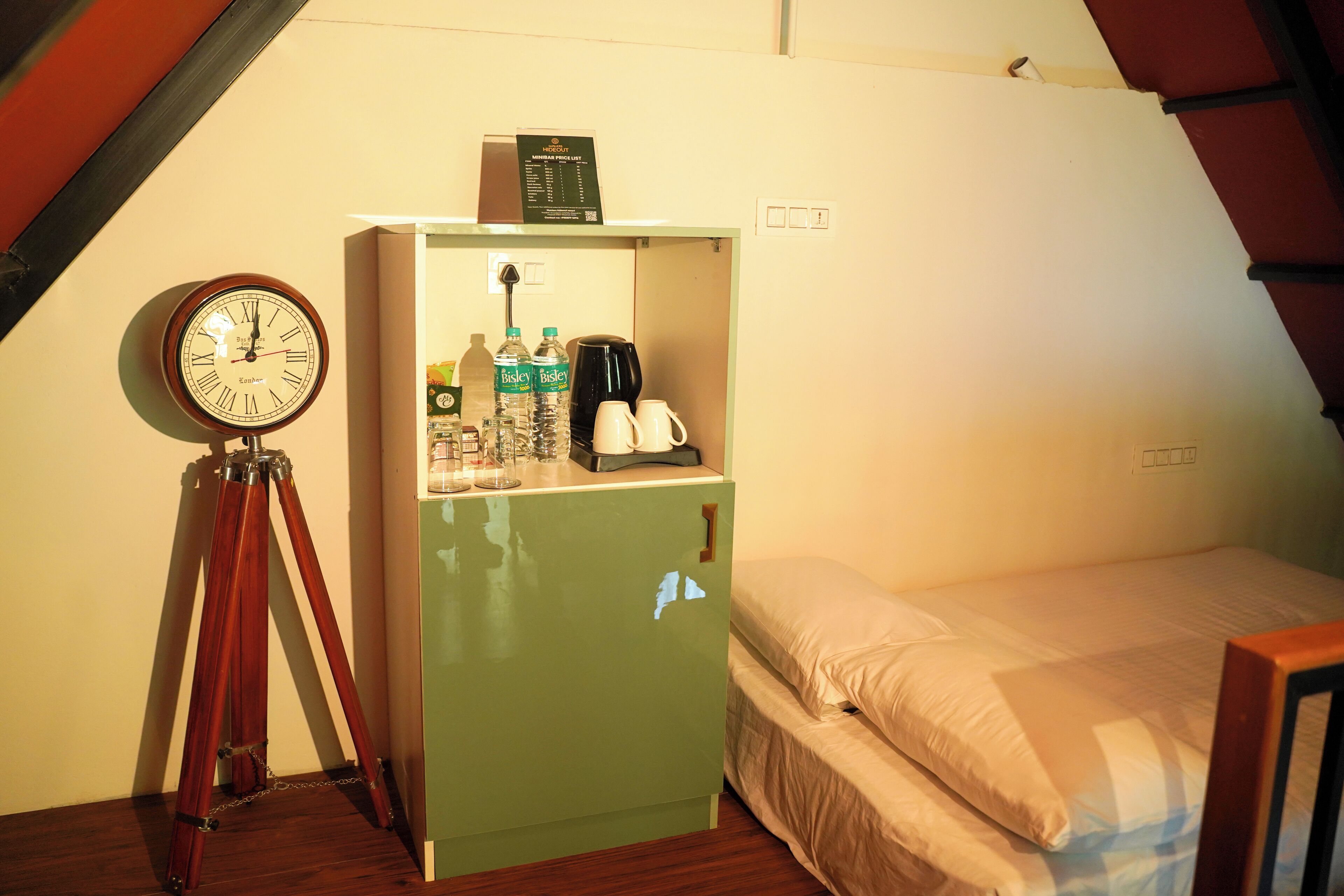 Una stanza piccola e verde con un letto, una sveglia di legno e uno scaffale con bottiglie e una macchina da caffè.