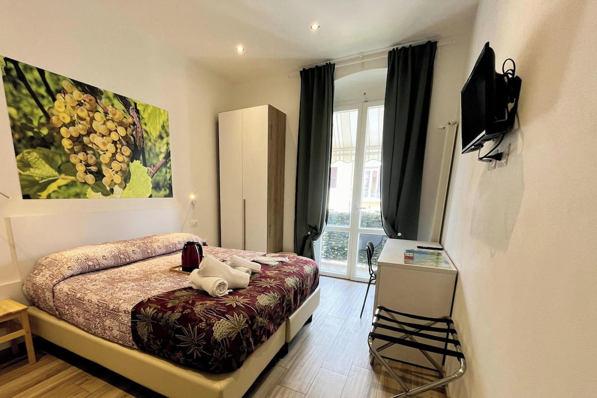 Quarto de hotel com cama, escrivaninha, cadeira, guarda-roupa, pintura e janela com cortinas.