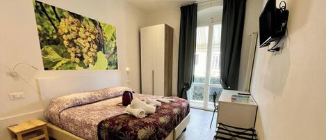 A hotel room with a bed, a desk, a chair, a wardrobe, a painting, and a window with curtains.