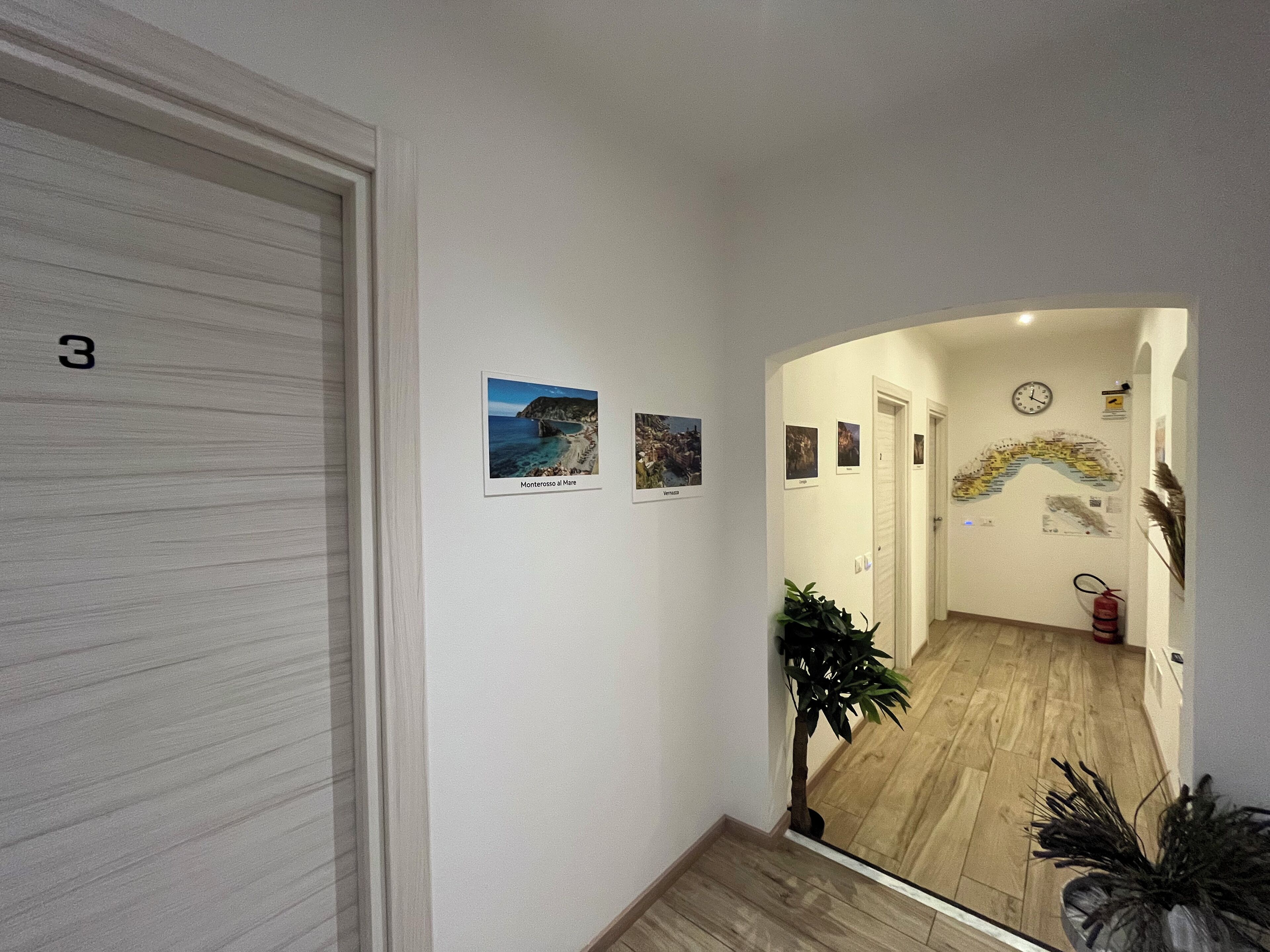 Un pasillo con piso de madera, una puerta con la etiqueta '3' y un espejo que refleja una habitación con un reloj y una planta.