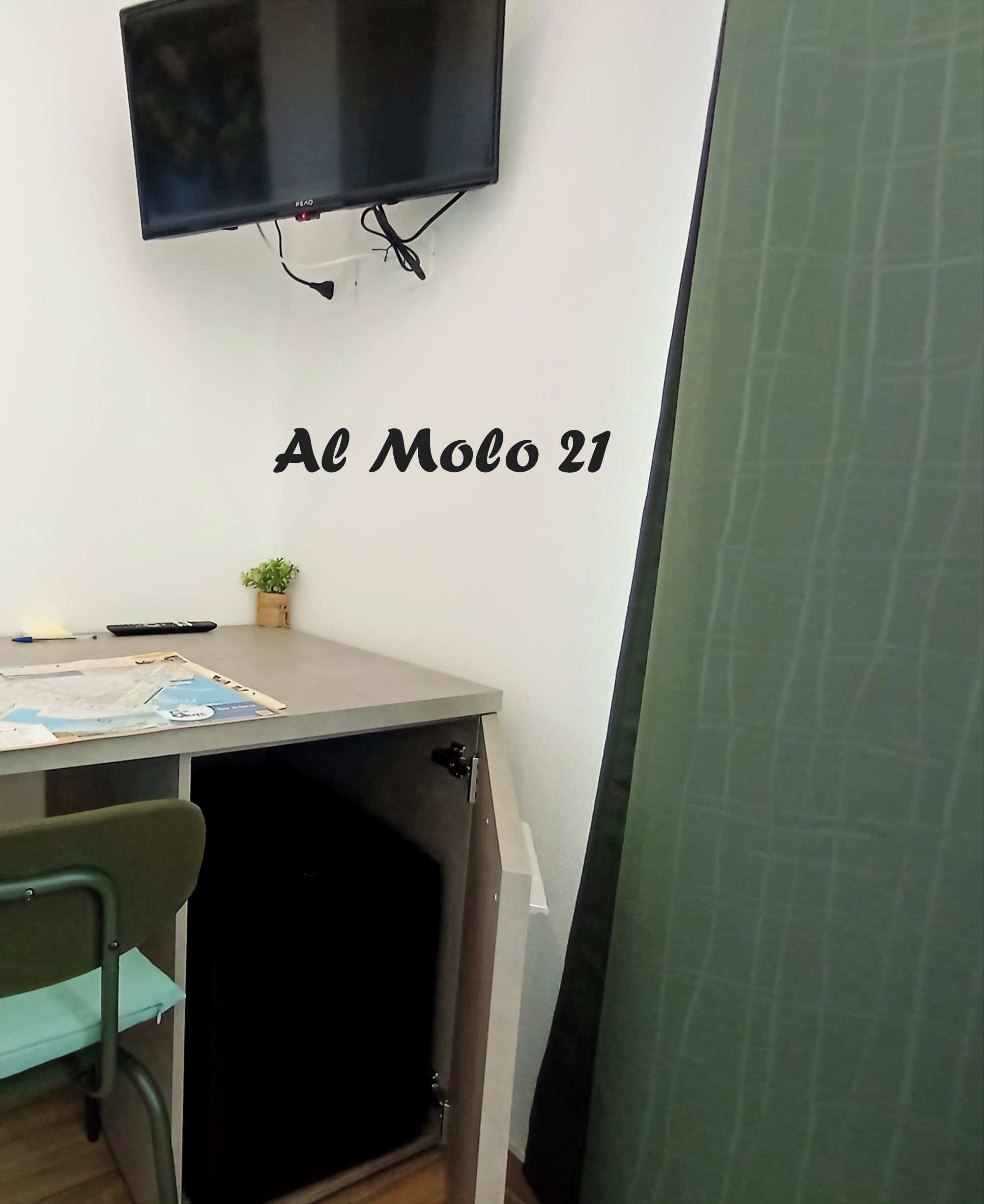 Quarto com escrivaninha, televisão na parede e placa com a inscrição 'Al Molo 21'.