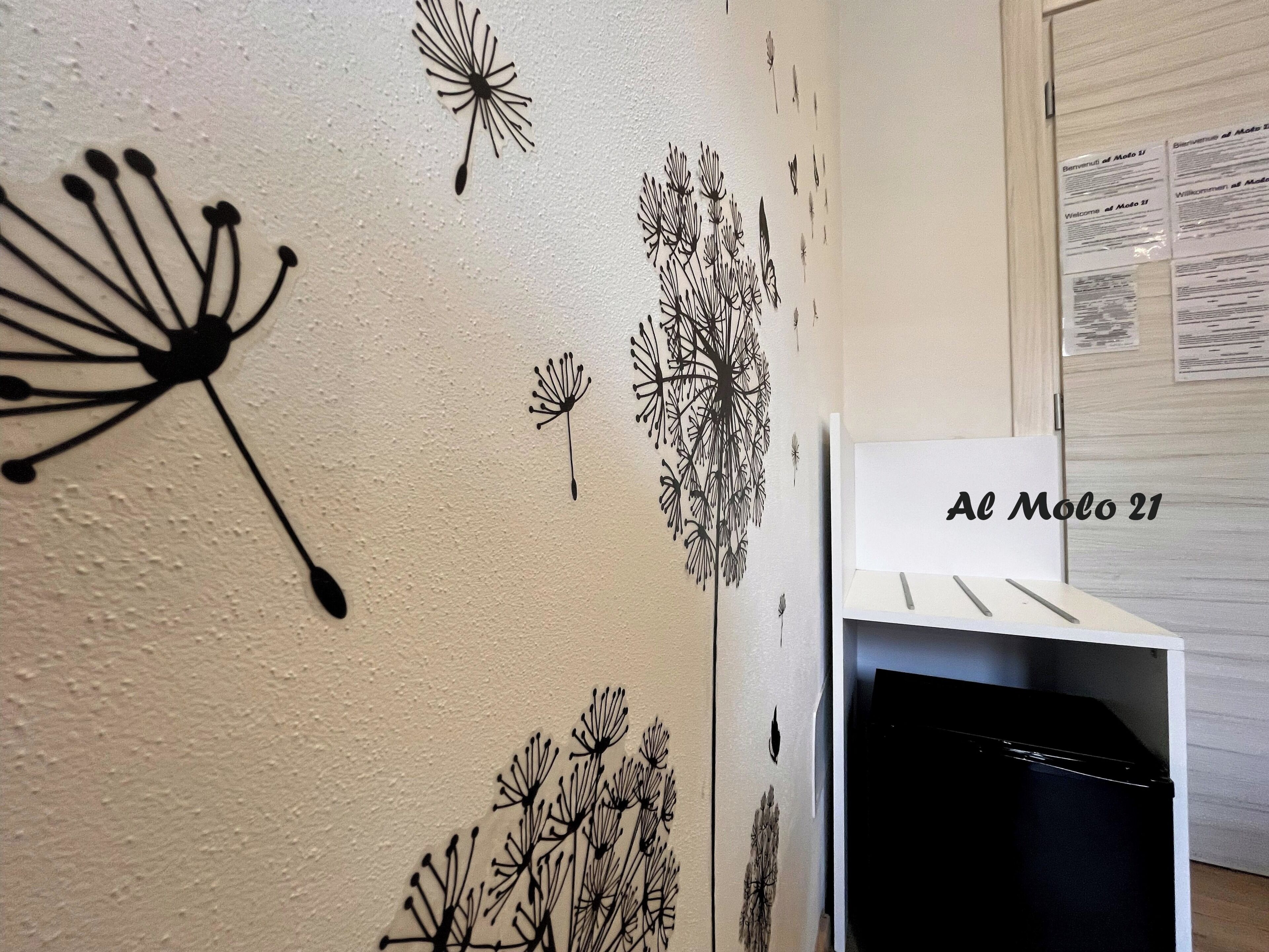 Eine Wand mit schwarzen Löwenzahn-Aufklebern, ein weißes Regal mit einem schwarzen Gegenstand und ein Schild mit der Aufschrift 'Al Moto 21'.