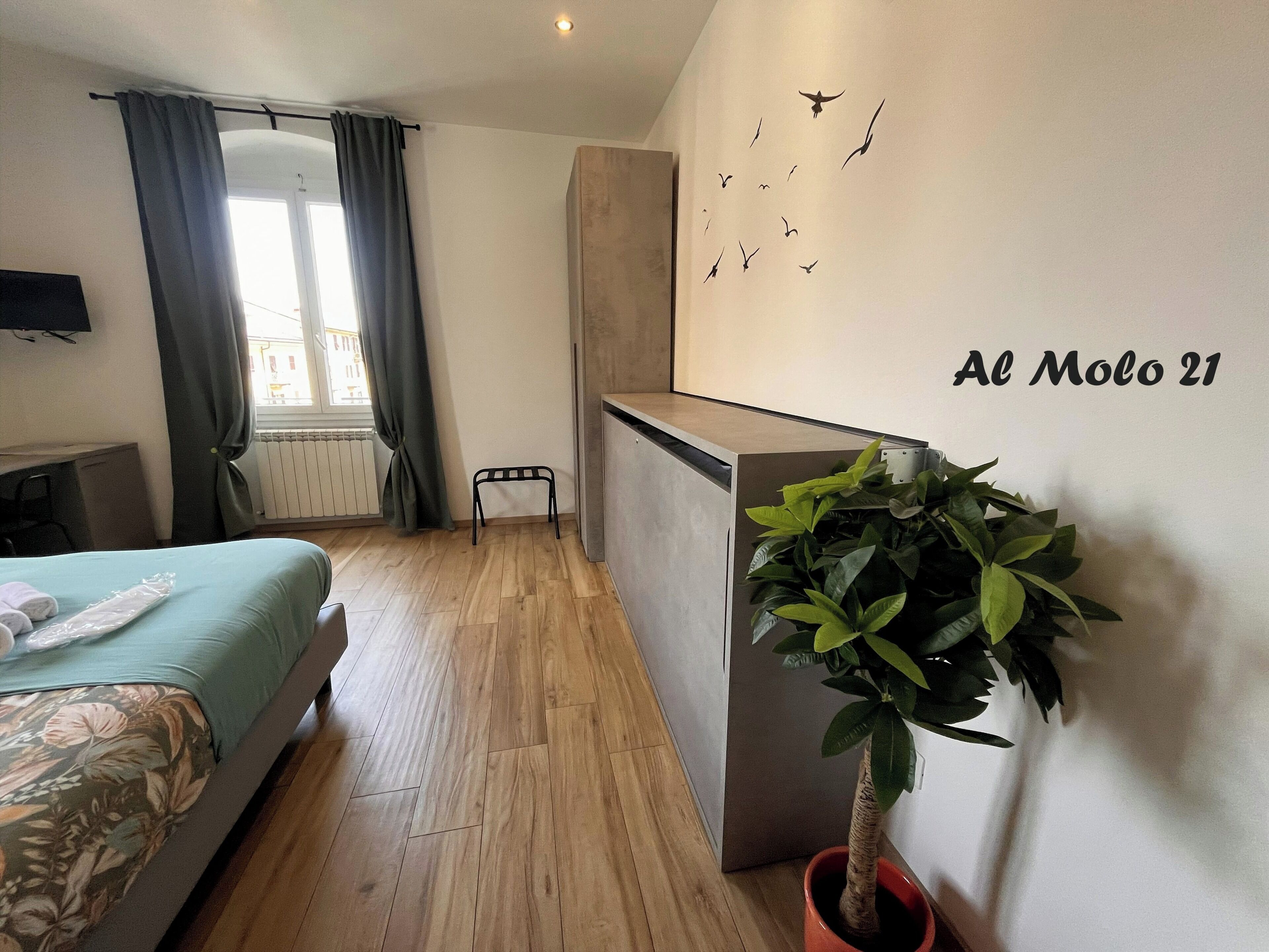 A hotel room with a bed, a small stool, a potted plant, a wall-mounted TV, and a window with curtains.