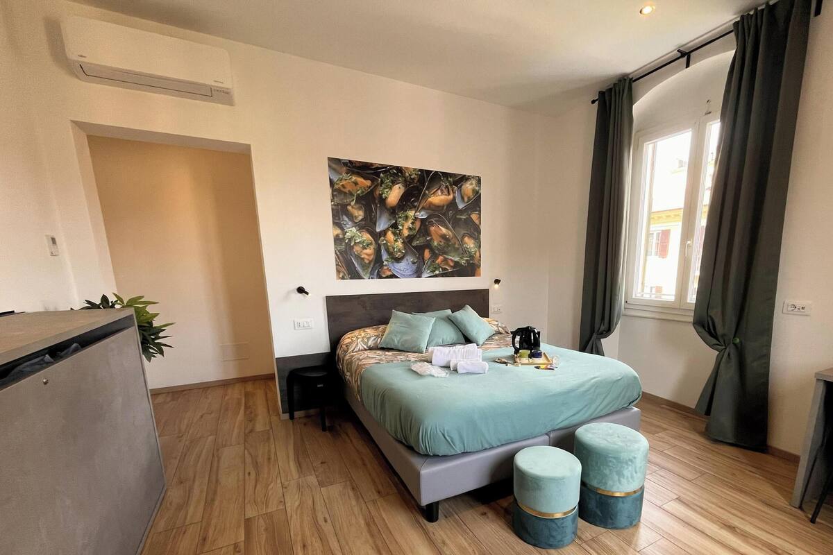 Quarto com cama, mesas de cabeceira, escrivaninha, janela com cortinas e ar-condicionado fixado na parede.