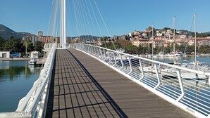 A pedestrian bridge with a wooden walkway and white railings, overlooking a marina with boats and a town in the background. - al molo 21 (La Spezia)