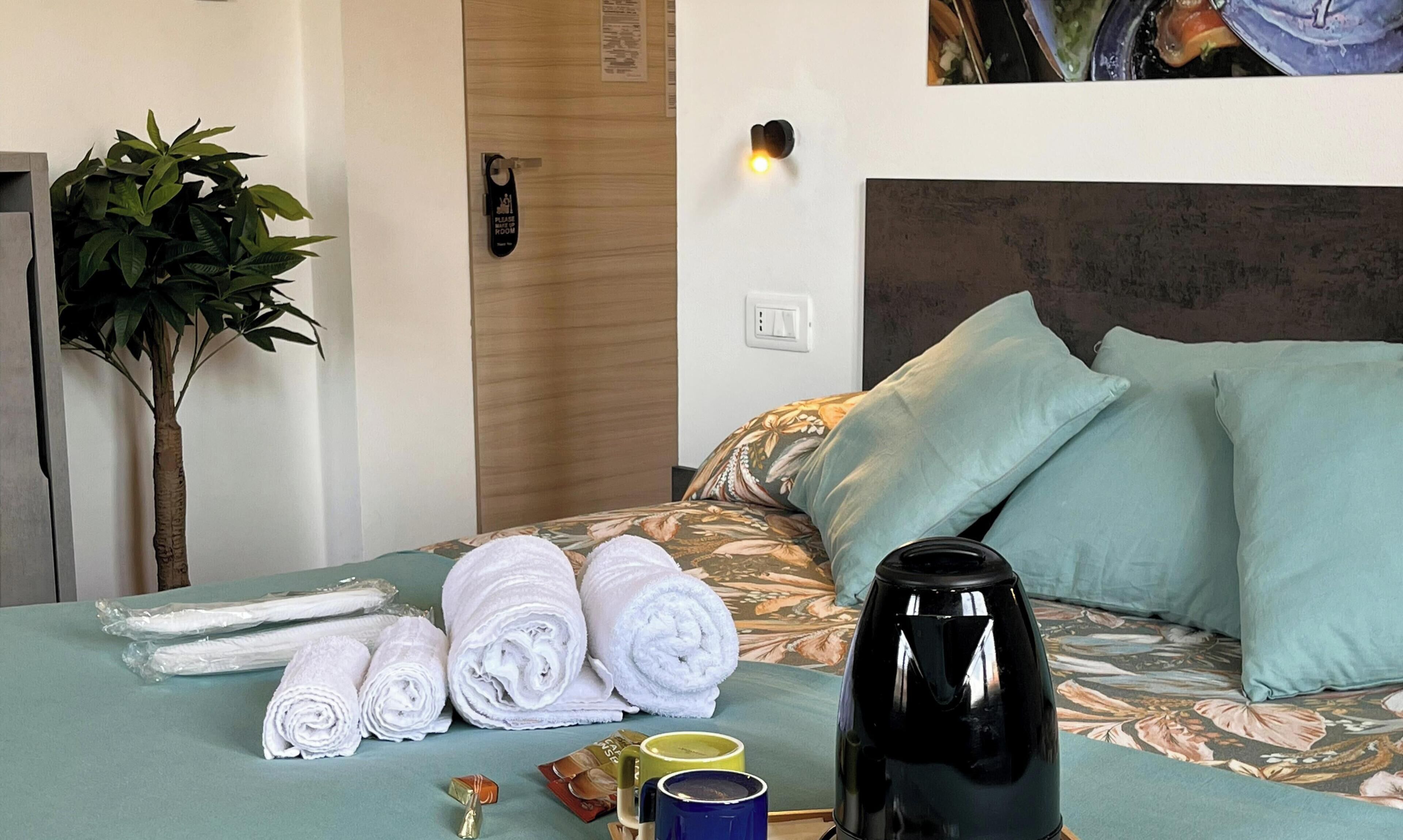 A hotel room with a bed, pillows, towels, a kettle, and a plant.