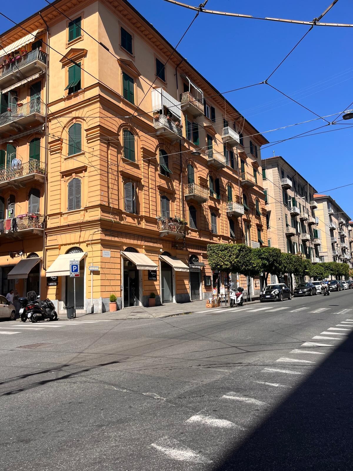 Une rangée d’immeubles à plusieurs étages dotés de balcons, un ciel dégagé et une rue bordée de voitures garées.