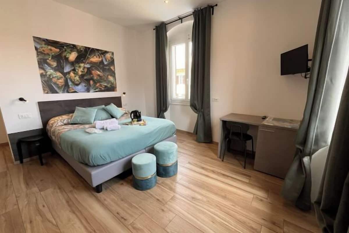 Quarto com cama, mesas de cabeceira, escrivaninha, cadeira, janela com cortinas e um quadro na parede.