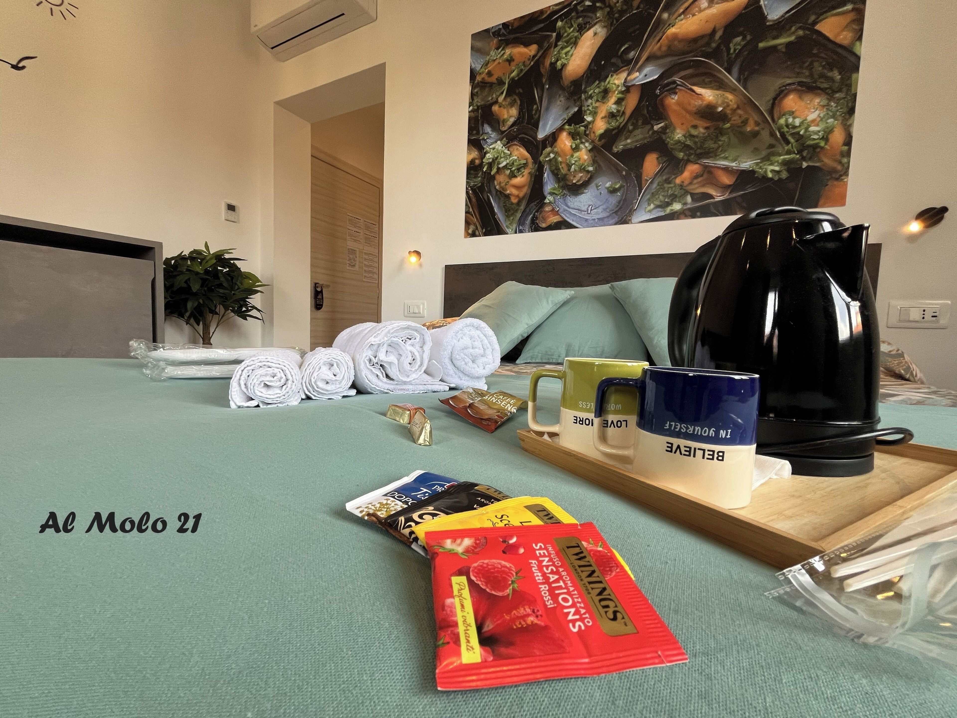 Een net opgemaakt bed met opgerolde handdoeken, een dienblad met mokken en snacks, een schilderij van kokkels, en een zwarte koffiezetapparaat.