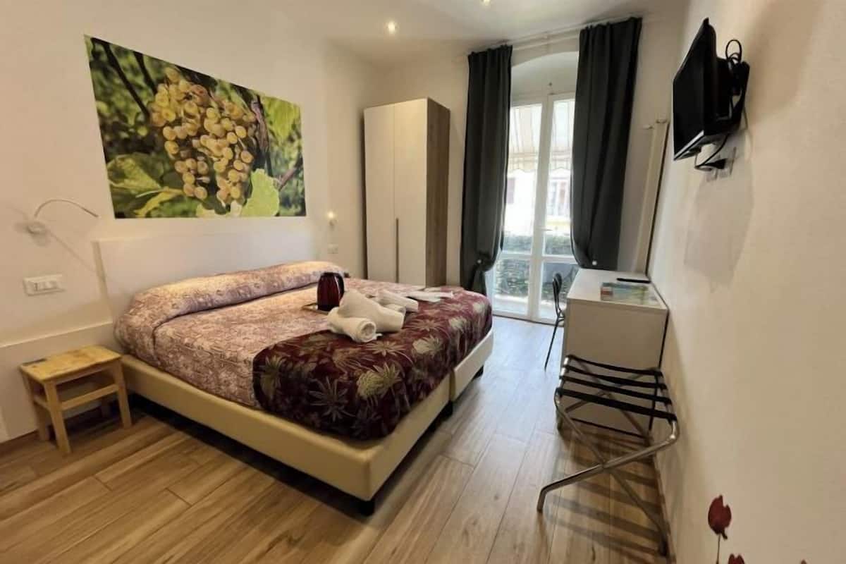 Quarto com cama, uma mesinha, guarda-roupa, escrivaninha e janela com cortinas.