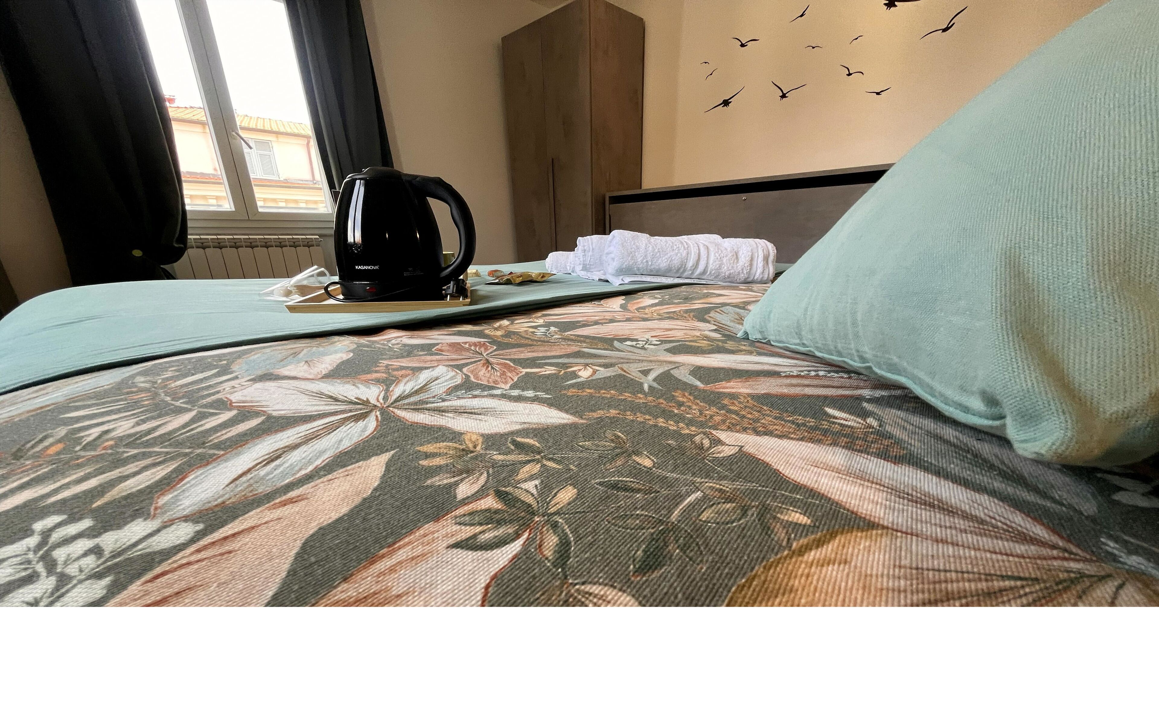 Un lit avec une literie à motifs floraux, une bouilloire et des serviettes sur une table de chevet.
