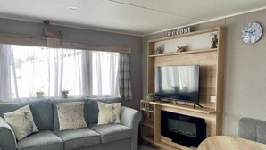 A living room with a grey sofa, a flat-screen TV, a wooden entertainment unit, and a fireplace. - Cosy 3 Bedroom Caravan in South Cerney (Cirencester)