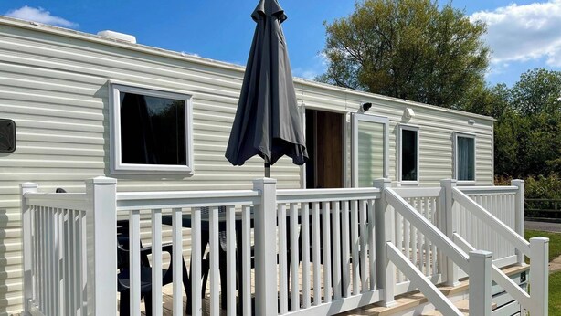 A mobile home with a white exterior, a deck with white railings, and a dark-colored umbrella. - Cosy 3 Bedroom Caravan in South Cerney (Cirencester)