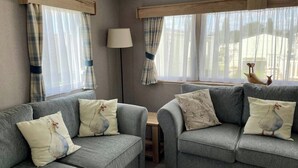A cozy living room with two grey sofas, each with a white pillow featuring a bird design. There is a wooden-framed window with sheer curtains, a floor lamp, and a view of a chicken outside the window. - Cosy 3 Bedroom Caravan in South Cerney (Cirencester)