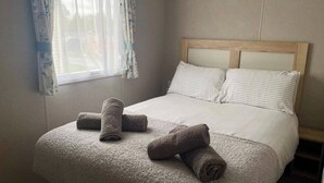 A neatly made bed with a headboard, a window with floral curtains, and rolled towels on the bed. - Cosy 3 Bedroom Caravan in South Cerney (Cirencester)