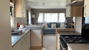 A compact kitchen with a stove, sink, and cabinets, leading to a seating area with a sofa and curtains. - Cosy 3 Bedroom Caravan in South Cerney (Cirencester)