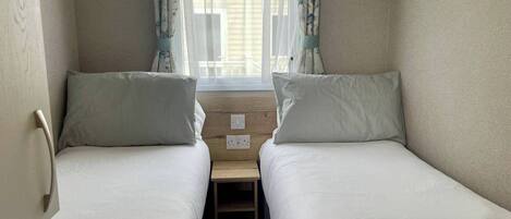 A small, cozy room with two single beds, a wooden nightstand, and a window with sheer curtains.