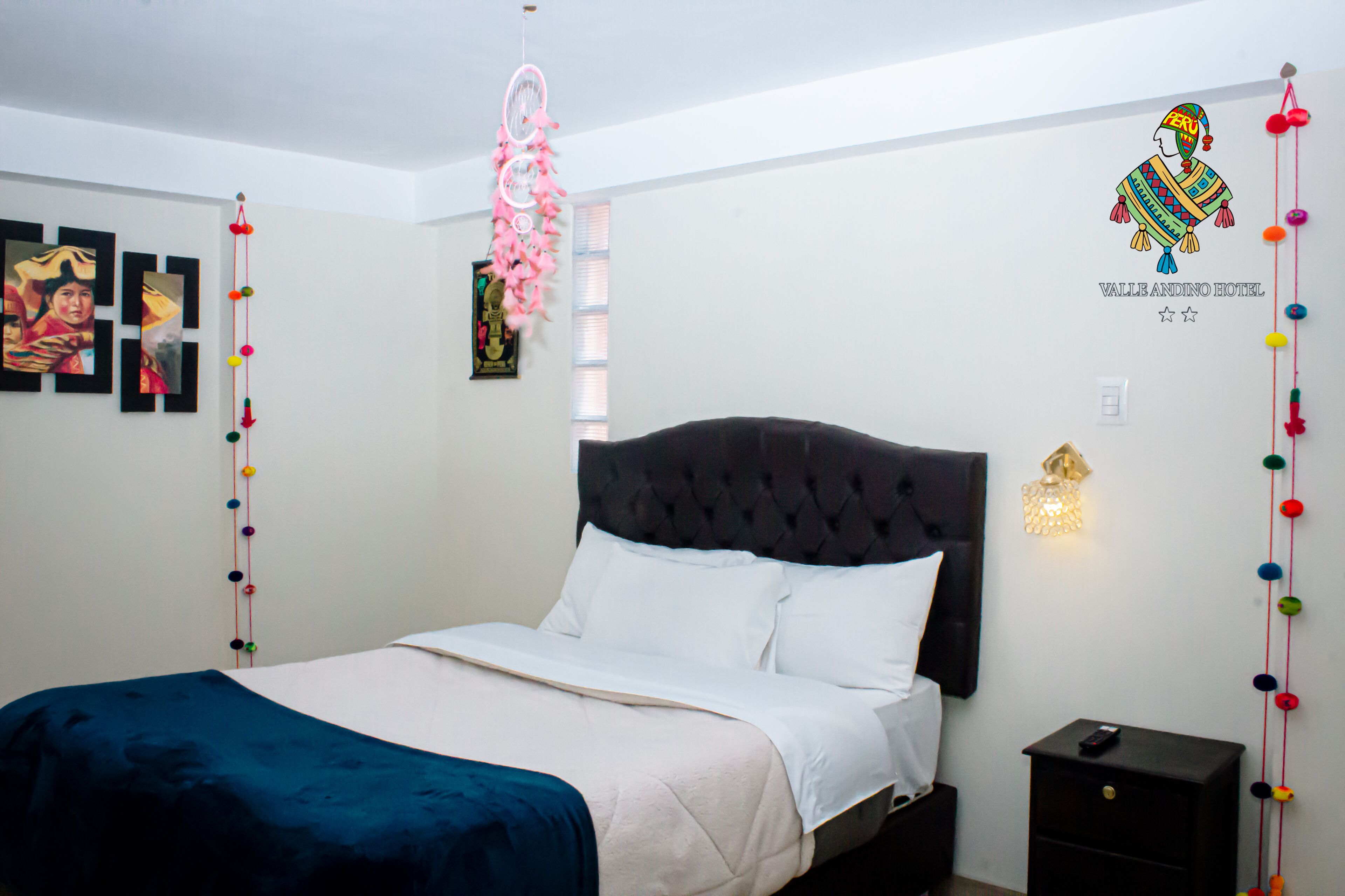 Una cama bien hecha con un cabecero oscuro, una mesita de noche con una lámpara y detalles decorativos en la pared, incluyendo cuadros enmarcados y un adhesivo mural de una figura tradicional.