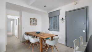 A dining area with a wooden table and white chairs, a clock on the wall, and a door with a window. - Modern Luxury Villa in Vlicha (4347+W3Q Lindos, Greece)