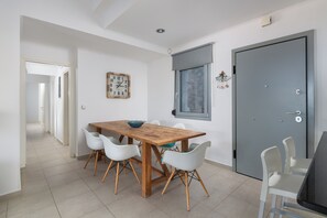 Uma sala de jantar com mesa de madeira e cadeiras brancas, um relógio na parede e uma porta com janela.