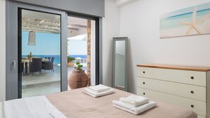 A hotel room with a bed, a dresser, a mirror, a painting, and a view of the sea through a glass door. - Modern Luxury Villa in Vlicha (4347+W3Q Lindos, Greece)