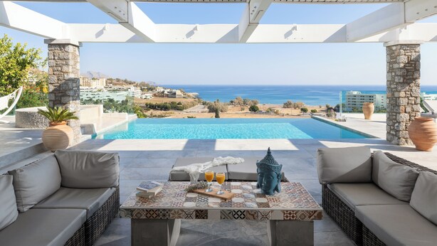 A poolside area with a view of the sea, a Buddha statue, and a table with two glasses of orange juice. - Modern Luxury Villa in Vlicha (4347+W3Q Lindos, Greece)