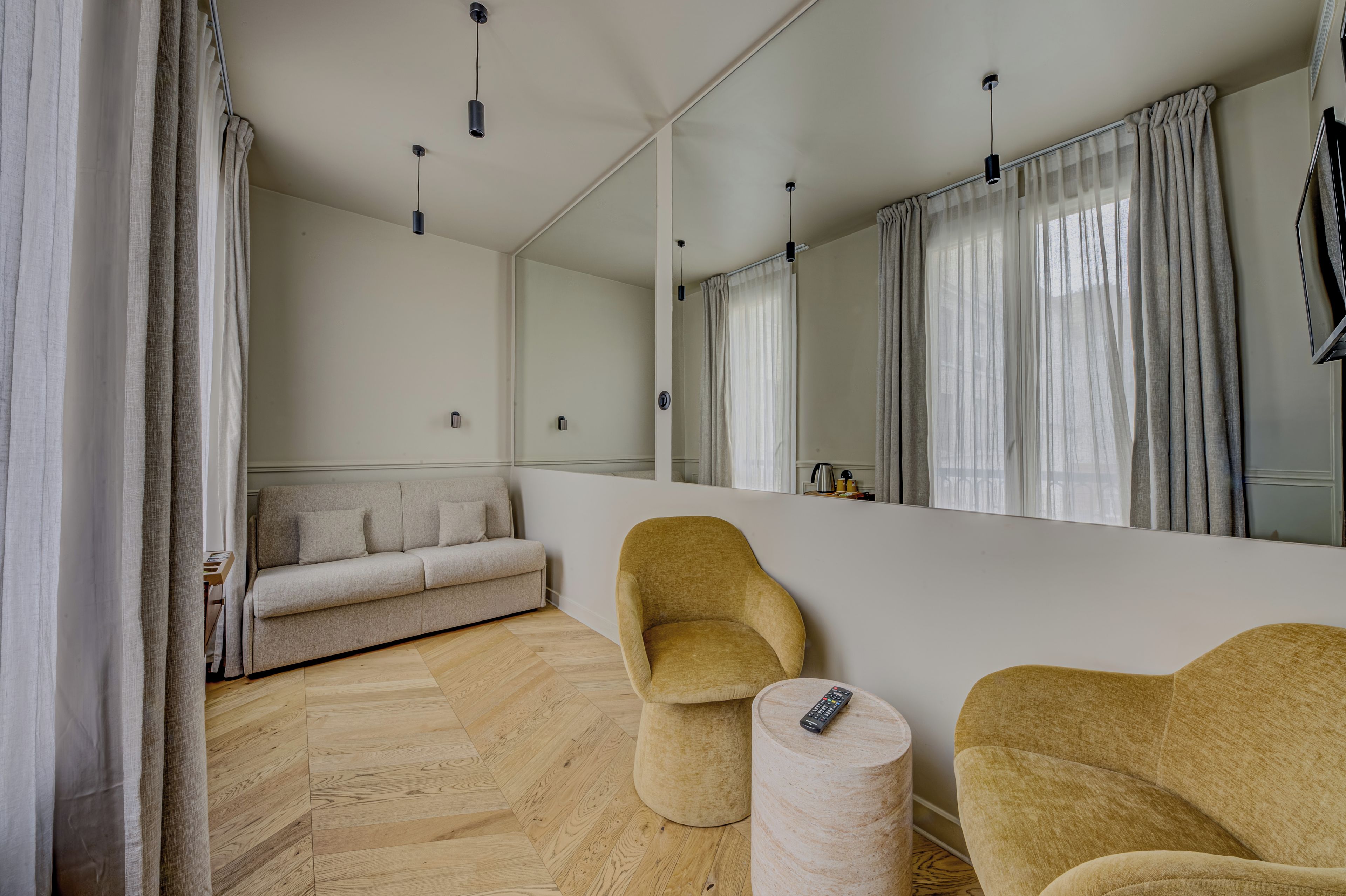Una habitación moderna con sofá, dos sillones, un taburete redondo y un espejo grande que refleja la habitación.