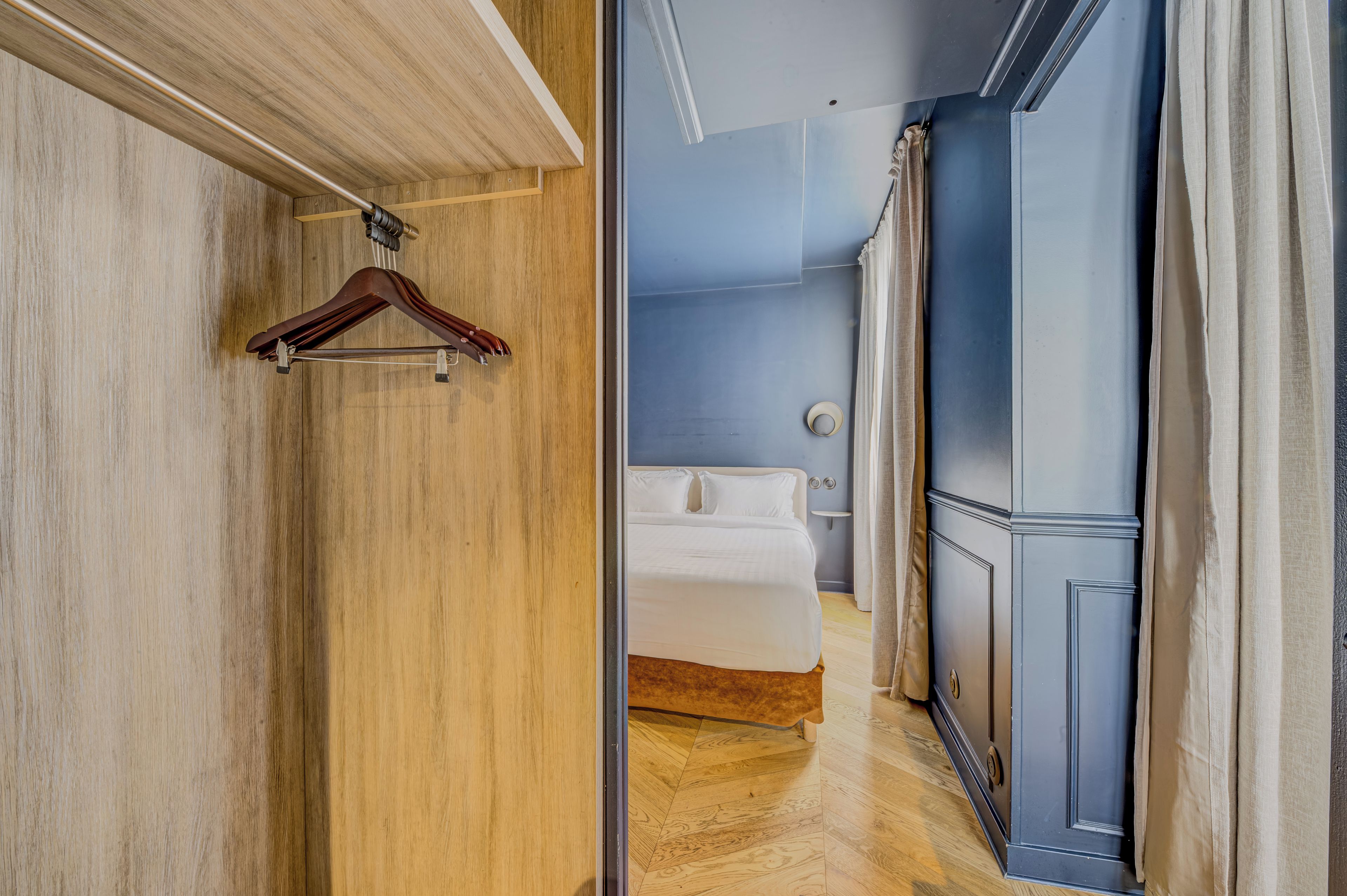 A hotel room with a wooden floor, a bed with white bedding, a wooden wardrobe, and a hanging garment rack.