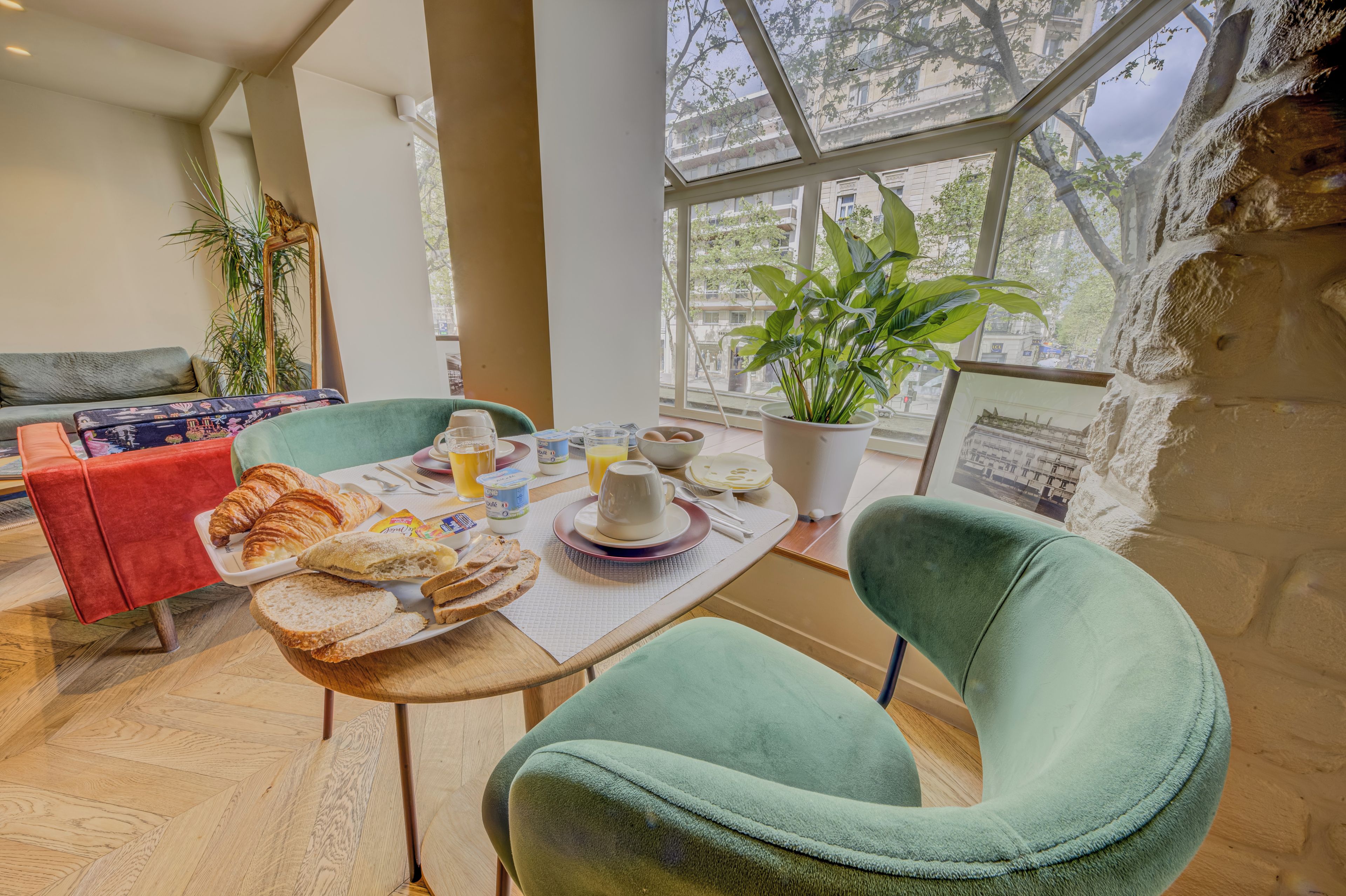 A breakfast setting with croissants, bread, yogurt, and juice on a table with a view of the city through large windows.