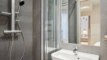 Un baño moderno con cabina de ducha de vidrio, un lavabo blanco y un espejo grande que refleja un dormitorio.