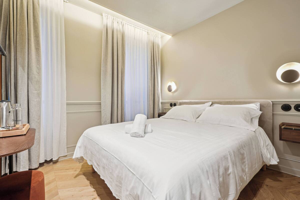 Een hotelkamer met een groot bed, een nachtkastje met een lamp en een raam met gordijnen.