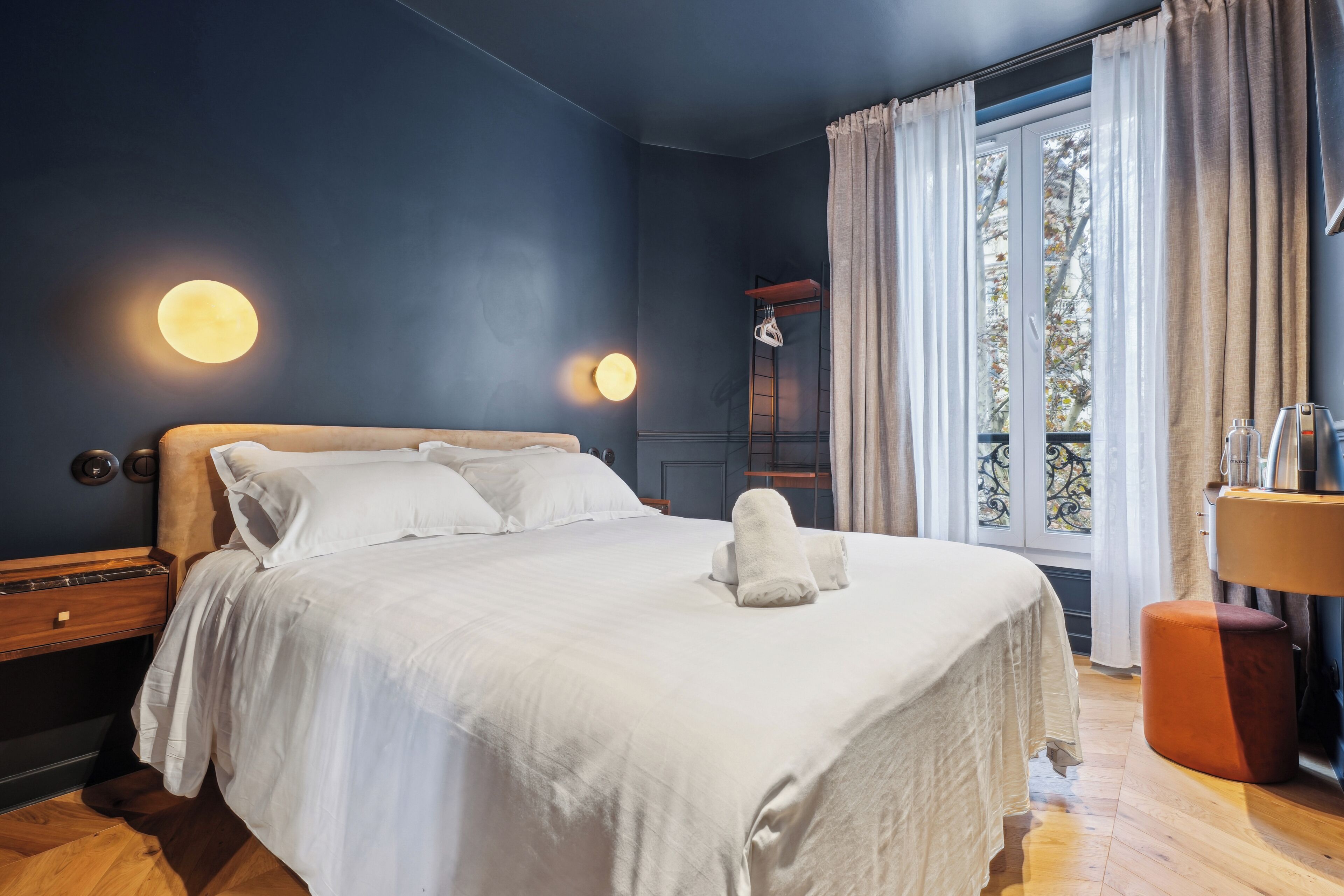 A neatly made bed with white linens and a folded towel, a bedside table with a lamp, and a window with curtains.