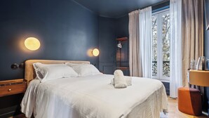 Un letto rifatto con lenzuola bianche e un asciugamano piegato, un comodino con una lampada e una finestra con tende.