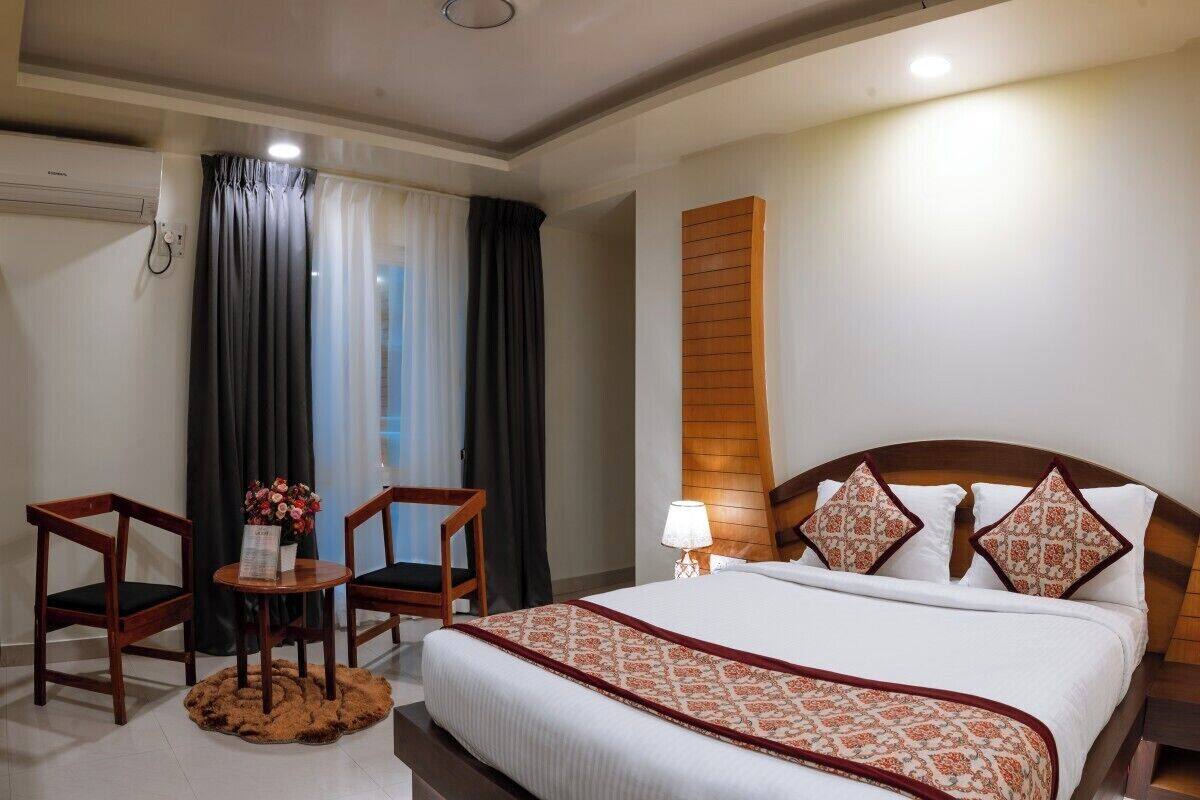 A hotel room with a bed, two wooden chairs, a small table with a vase of flowers, a lamp, and a window with curtains.