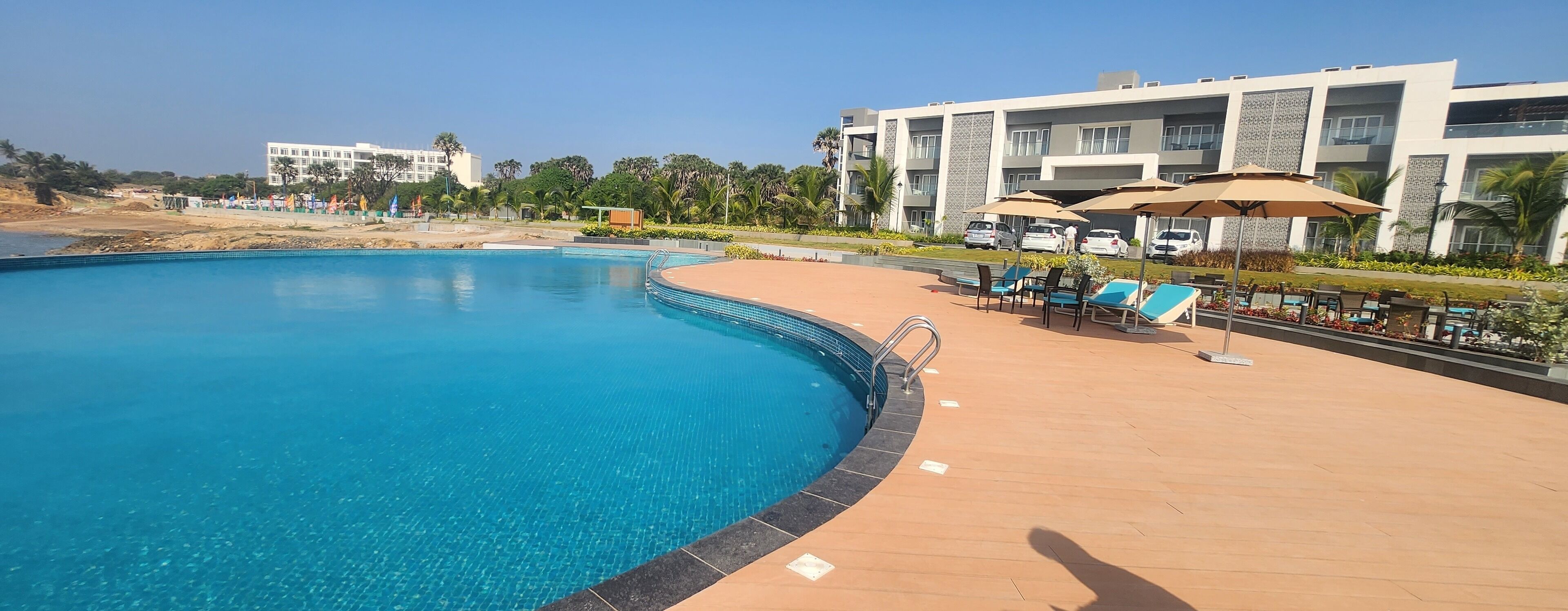 A swimming pool with a curved edge, surrounded by a tiled deck. There are lounge chairs and umbrellas, and a building with multiple floors and balconies in the background.