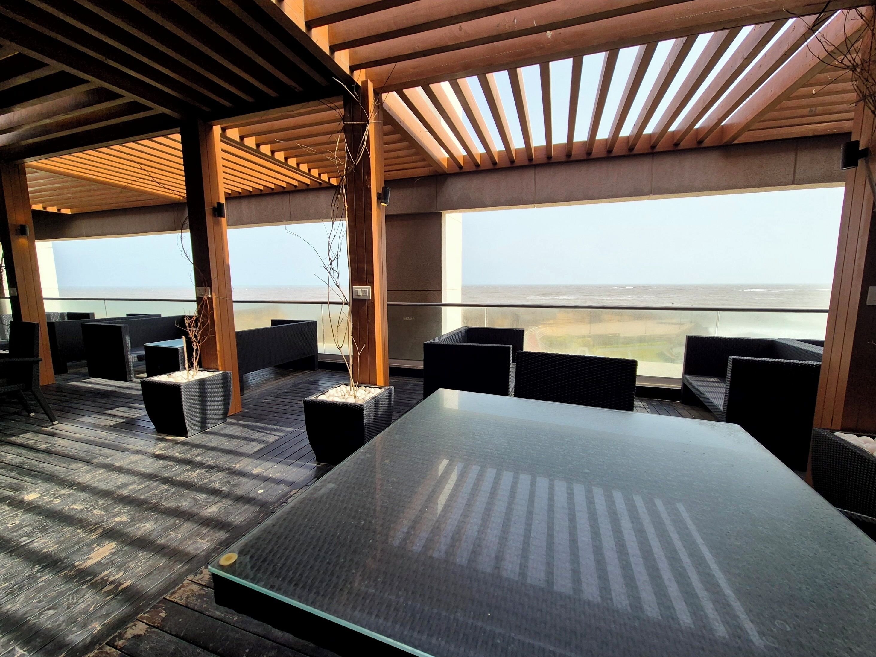 Un área de estar al aire libre con una mesa grande y sillas, vigas de madera y vista al océano.