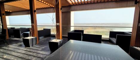 An outdoor seating area with a large table and chairs, wooden beams, and a view of the ocean.