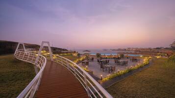 A wooden deck with a railing, outdoor seating area with tables and chairs, and a pool in the background.