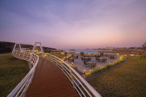 A wooden deck with a railing, outdoor seating area with tables and chairs, and a pool in the background. - Ginger Diu Jalandhar Beach (Diu)