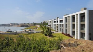 A modern hotel with a waterfront view, featuring a long building with balconies, a landscaped garden, and a pathway leading to a dock. - Ginger Diu Jalandhar Beach (Diu)