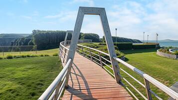 Un puente peatonal de madera con barandillas de metal, que lleva a un área ajardinada con un mirador y vista al mar.