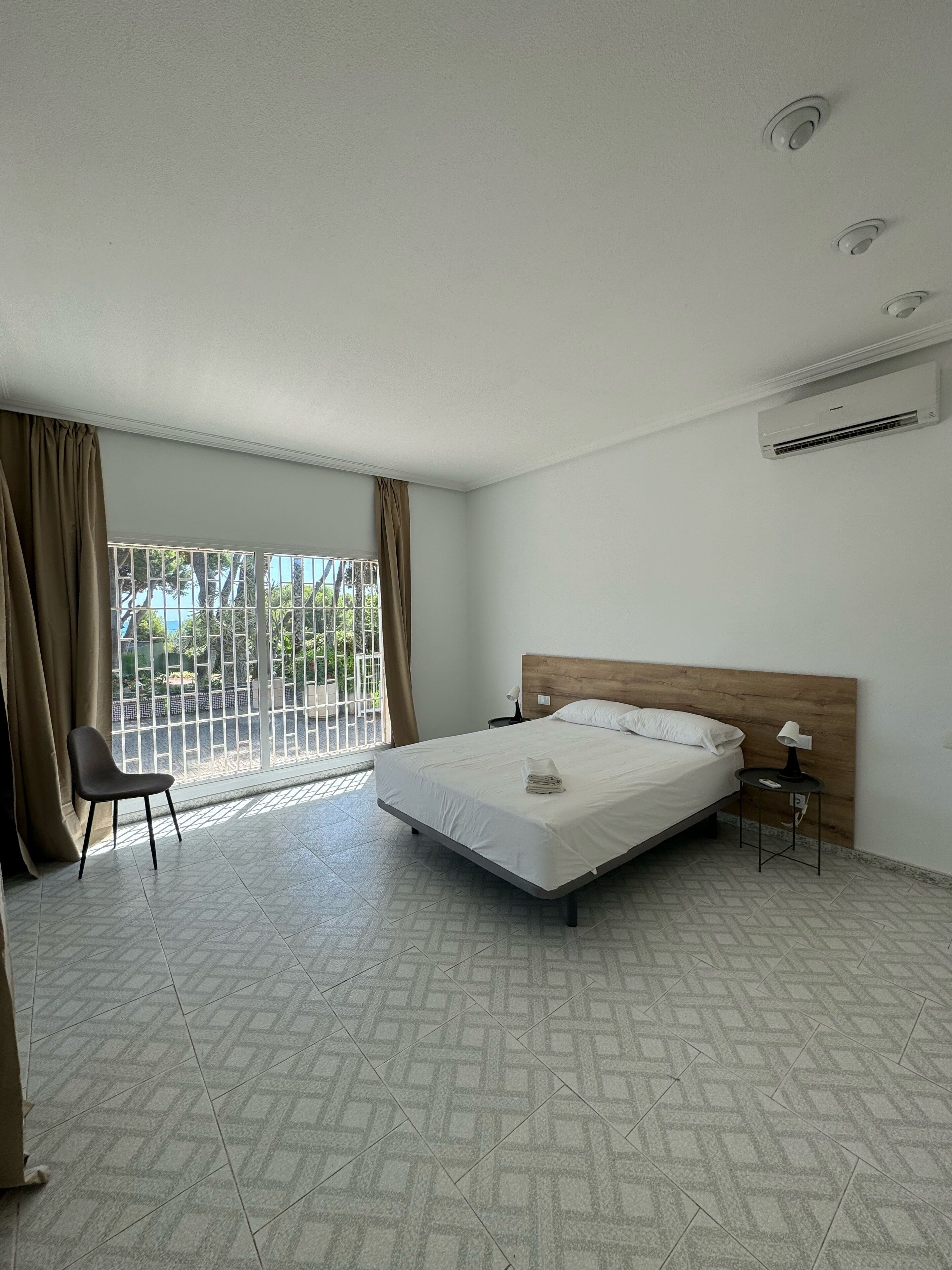Ein Hotelzimmer mit Bett, Sessel, Nachttisch, Klimaanlage und Blick ins Freie durch ein Fenster mit Vorhängen.
