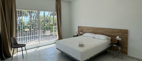 Ein Hotelzimmer mit Bett, Sessel, Nachttisch, Klimaanlage und Blick ins Freie durch ein Fenster mit Vorhängen.
