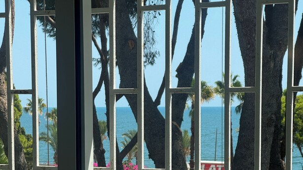 Blick durch ein Fenster auf einen Pool und Palmen, mit freier Sicht auf den Ozean und ein Gebäude im Hintergrund.