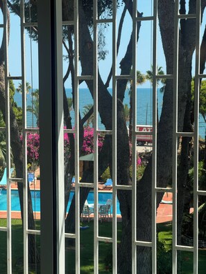 A view through a window with a pool and palm trees, with a clear view of the ocean and a building in the background. - Villa Marinada (Alcanar)