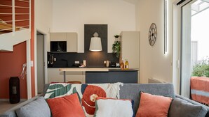 Ein modernes Wohnzimmer mit grauem Sofa, bunten Kissen, einer Küche im Hintergrund und einem großen Fenster.
