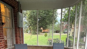 A covered patio with two chairs and a small table, a brick wall, and a view of a green lawn and trees. - Spacious 4-Bdrm house in Ruston with WiFi and AC & Wonderful front & back porch (Ruston)